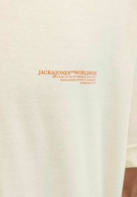 Šviesiai smėlio spalvos medvilninis marškinėliai su oranžiniu spaudiniu, kuriame parašyta "JACK & JONES WORLDWIDE", ir papildomi prekės ženklo detalės žemiau.