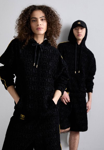 Felpe con cappuccio nere caratterizzate da un motivo testurizzato "VERSACE", arricchite da strisce gialle. Modello in pantaloni corti che mostra un design coordinato.