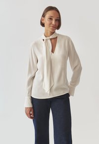 White blouse with a tied neck scarf, long sleeves, and a keyhole cutout at the chest. Worn with dark blue denim jeans. Smooth fabric texture.