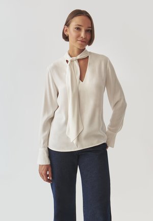 White blouse with a tied neck scarf, long sleeves, and a keyhole cutout at the chest. Worn with dark blue denim jeans. Smooth fabric texture.