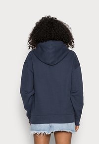 Felpa con cappuccio di colore blu navy, dal fit rilassato, con polsini e orlo a coste e texture morbida; indossata con pantaloni shorts in denim chiaro strappati.