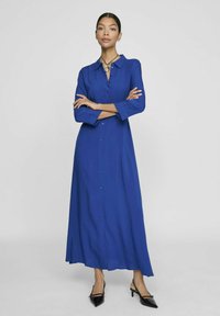 Mėlyna ilga marškinėlių suknelė, pagaminta iš plaukioto audinio, su mygtukais priekyje, trijų ketvirčių rankovėmis ir tiesia siluetu. Dėvima su juodais aukštakulniais.