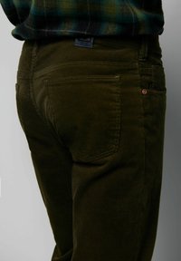 Donkergroene corduroy broek met verticale ribbels, voorzien van twee achterzakken en een kleine rode accent bij de zak. Textuur van de stof zichtbaar.