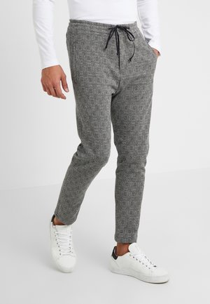 Mann trägt graue karierte Kordelhose und weiße Sneakers, steht mit einer Hand in der Tasche vor einem einfarbigen Hintergrund.