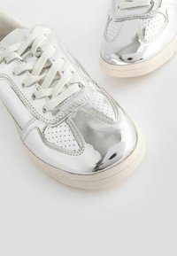 Sneaker argentati e lucidi con lacci bianchi, punta perforata e suola in gomma beige. Presentano una superficie liscia e riflettente.