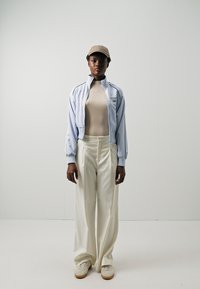 Chaqueta de cremallera azul claro sobre una camiseta ajustada beige, combinada con pantalones anchos blancos y zapatillas color crema. Una gorra beige completa el look.