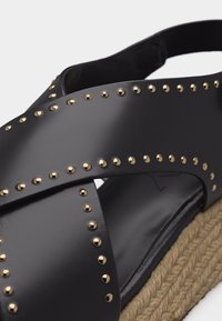 Sandale en cuir noir avec des lanières croisées ornées de clous dorés et une semelle en fibres naturelles tressées.