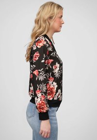 Veste bomber noire à motifs floraux avec des fleurs rouges et blanches, poignets et taille côtelés, associée à un jean en denim bleu clair.