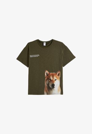 Oliven grøn bomulds t-shirt med et stort Shiba Inu hundegrafik på forsiden og hvid tekst ovenfor. Rund hals med korte ærmer.