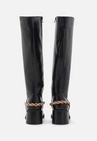 Botas negras de imitación piel hasta la rodilla con un tacón bloque grueso y acentos de cadena metálica trenzada en el tobillo. Cierre de cremallera en la parte posterior.