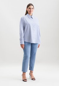Gestreifte Button-Up-Bluse in Blau und Weiß; lockere Passform; umgeschlagene Ärmel. Kombiniert mit hellblauen Denim-Jeans und schwarzen Absatzsandalen.
