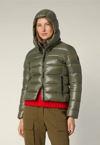 Giacca imbottita verde oliva con cappuccio aderente, chiusura con zip e tasche laterali. Indossata sopra un maglione di lana rosso e pantaloni khaki.