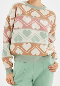 Person som bär en pastellfärgad tröja med hjärtformade mönster i ljusgrön, rosa och beige, kombinerat med ljusgröna högmidjade byxor.