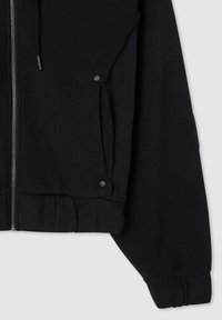 Felpa nera con zip, pannelli laterali a costine, bottoni a pressione sulle tasche, vita elasticizzata e maniche lunghe con polsini, su uno sfondo chiaro.