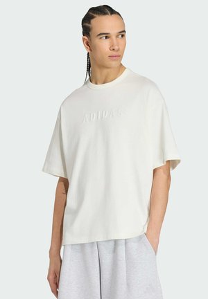 Młody mężczyzna z zaplecionymi włosami, ubrany w oversize'ową białą koszulkę Adidas i jasno szare spodenki, patrzący na bok.
