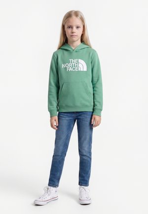 Dieťa s dlhými blond vlasmi, oblečené v zelenej mikine The North Face, modré džínsy a biele vysoké tenisky stojace na bielom pozadí.