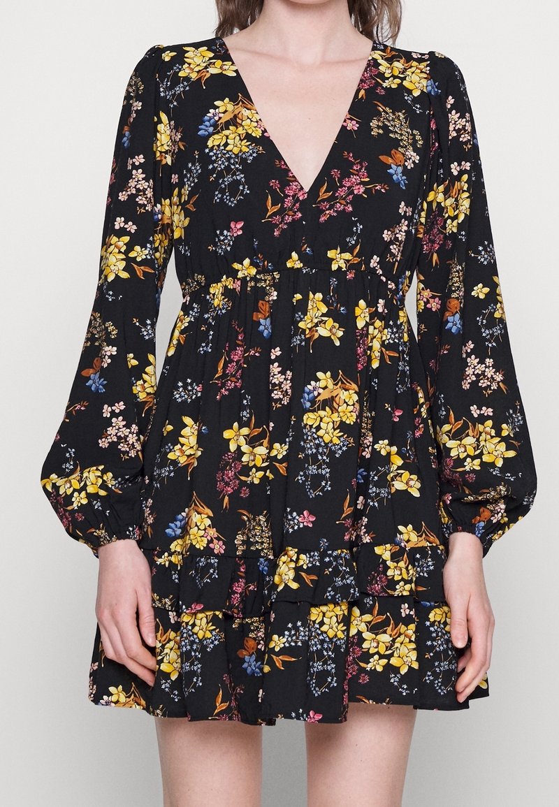 Vestido negro con estampado floral, con escote en V profundo, mangas largas abullonadas y dobladillo con volantes en capas. Presenta patrones florales en amarillo, rosa y azul sobre un fondo oscuro.