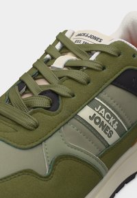 Κοντινή λήψη πράσινου και μπεζ αθλητικού παπουτσιού Jack & Jones με υφασμάτινες και σουέντ υφές, δεμένους κορδόνια και λογότυπο της μάρκας στο πλάι και στη γλώσσα.