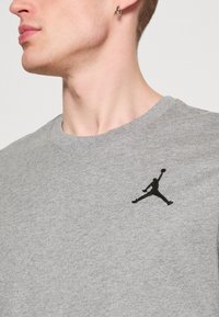 Tricou din bumbac gri cu guler rotund, având un logo Jumpman brodat negru în zona stângă a pieptului. Textură netedă, design casual.