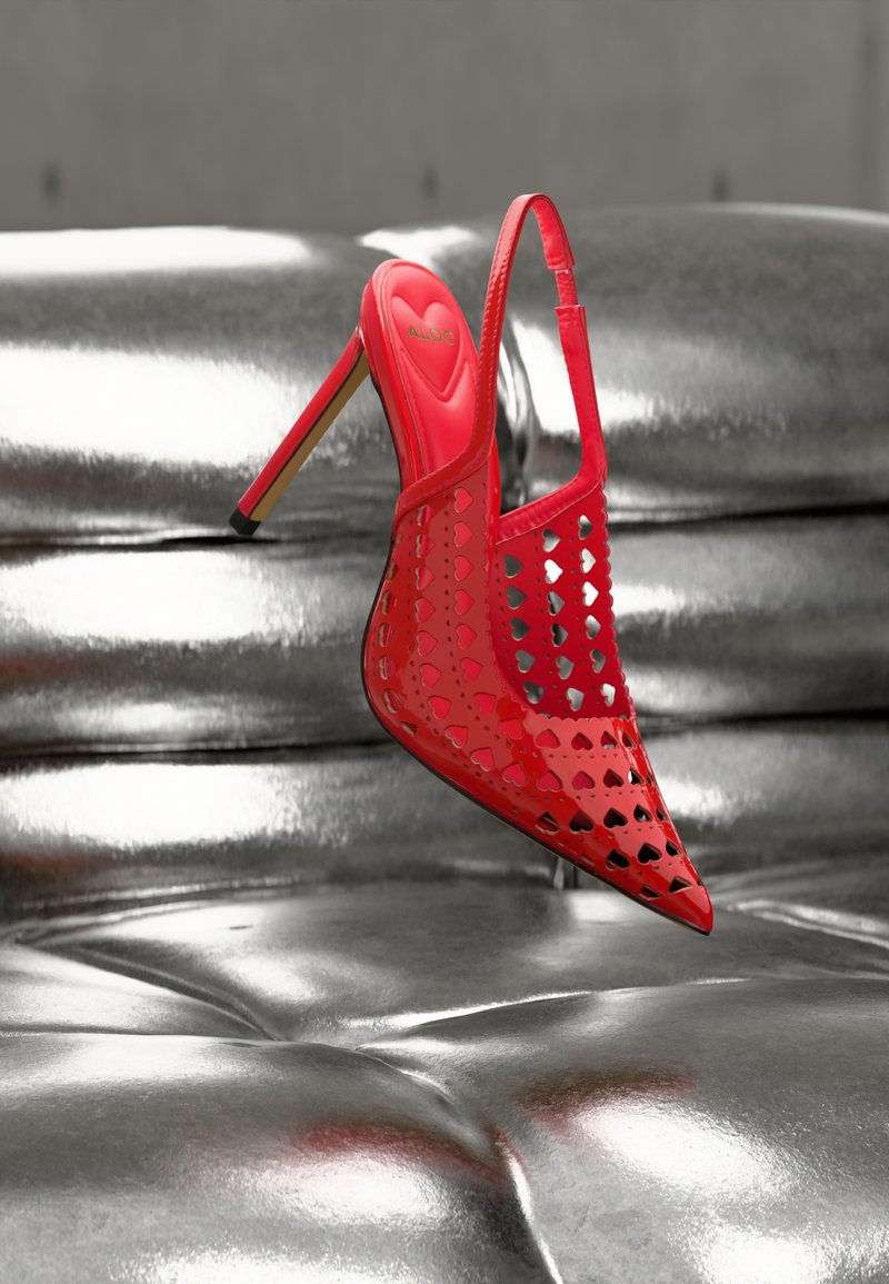 Chaussure rouge à talons hauts avec bride arrière et découpes en forme de cœur, présentée sur une surface argentée brillante et capitonnée.