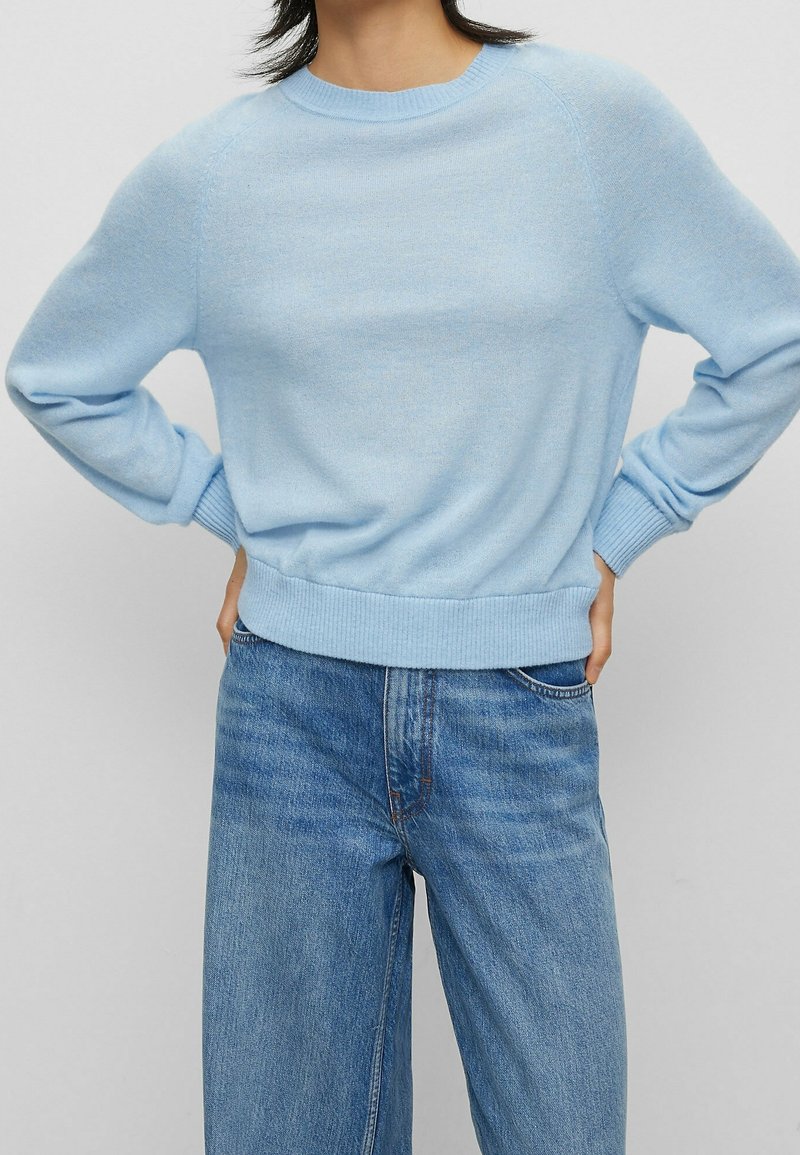 Personne portant un pull à manches longues bleu clair et un jean en denim bleu, les mains reposant sur les hanches, devant un fond uni.