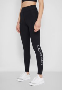 Černé legíny s vysokým pasem a bílým logem "CALVIN KLEIN" na levé noze. Hladký materiál, pružný střih, v kombinaci s bílými botami.