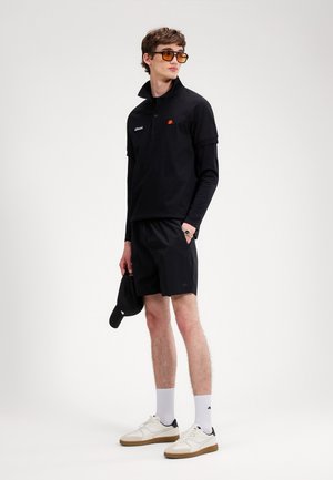 Bărbat tânăr purtând un polo negru cu straturi și mâneci lungi, pantaloni scurți negri, șosete albe, adidași albi, ținând o șapcă neagră, privind în lateral.