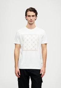 Bílý bavlněný tričko s čtvercovým designem v béžové barvě a kulatými vzory. Krátké rukávy a kulatý výstřih. Pod ním jsou nošeny černé kalhoty.