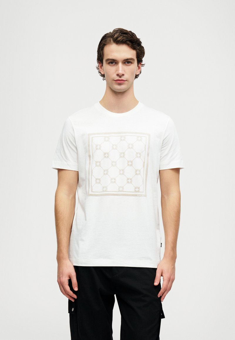 Bílý bavlněný tričko s čtvercovým designem v béžové barvě a kulatými vzory. Krátké rukávy a kulatý výstřih. Pod ním jsou nošeny černé kalhoty.