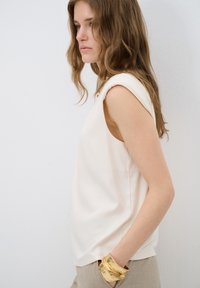 Blusa sin mangas de color crema con una textura suave, escote redondo y detalle de hombro enrollado. Combinada con una pulsera de brazalete dorada.