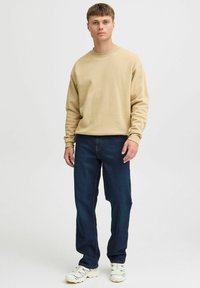 Sudadera beige en mezcla de algodón, de manga larga, con cuello redondo; combinada con unos vaqueros de mezclilla azul oscuro y zapatillas blancas con acentos azules.