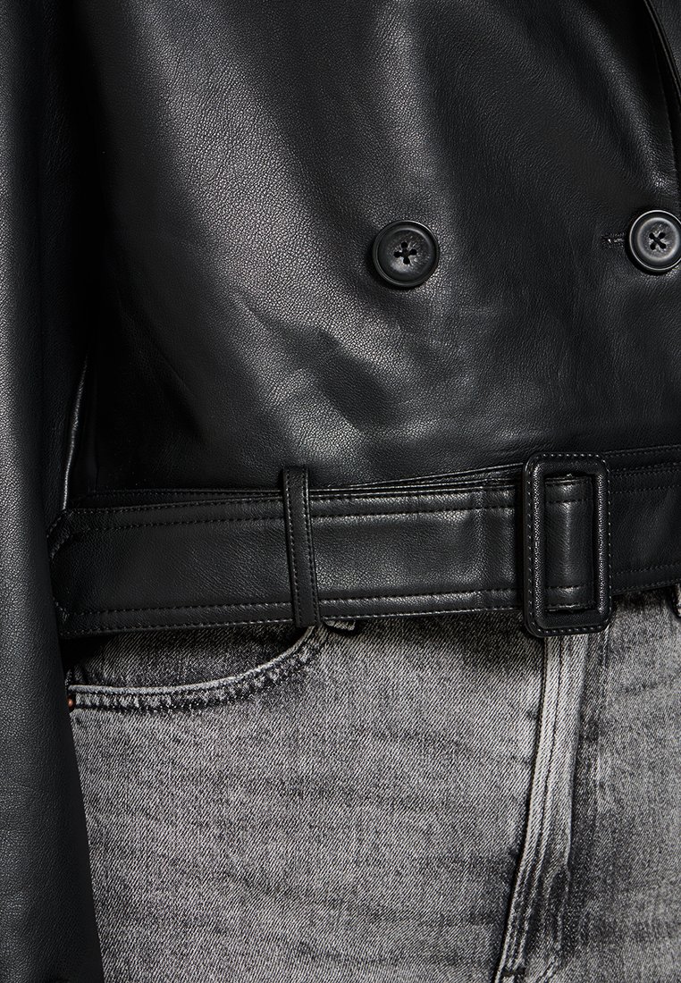Schwarze Lederjacke mit doppelreihigem Design, auffälligen Knöpfen und tailliertem Bund, kombiniert mit grauen Jeans.