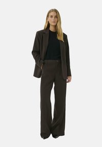 Bruine op maat gemaakte blazer over een zwarte trui, gecombineerd met bijpassende wijde broek. Smeuig materiaal met strakke lijnen en minimale hardware details.