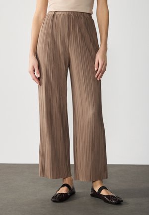 Frau trägt braune, hoch taillierte, weit geschnittene, plissierte Hosen, kombiniert mit schwarzen Ballettschuhen mit elastischen Riemen, steht auf grauem Boden.