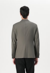 Chaqueta de traje gris con un diseño estructurado, solapas de pico y mangas largas con detalles de botones, exhibida desde la parte de atrás sobre un fondo neutro.