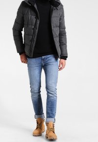 Chaqueta acolchada gris con capucha, suéter negro de cuello alto, jeans azul claro y botas tan con cordones. La chaqueta tiene botones y un tejido texturizado.