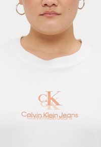 Bela pamučna majica s teksturiranim logotipom u ružičastom zlatu koji glasi "CK" i "Calvin Klein Jeans" ispod, s klasičnim okruglim izrezom.