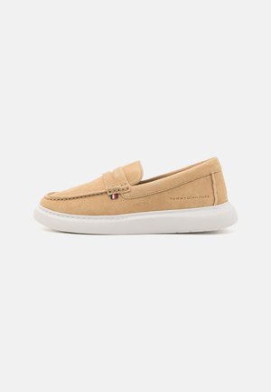 Beige suède instappers met een witte rubberen zool, voorzien van decoratieve stiksels en een klein logo-accent aan de zijkant.
