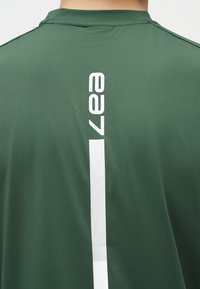 Πίσω όψη ατόμου που φοράει σκούρο πράσινο πουκάμισο με λευκά κάθετα γράμματα "ea7" και λευκή κάθετη ρίγα στο κέντρο.