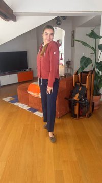 Γυναίκα που χαμογελά σε σαλόνι, φορώντας ένα κόκκινο φούτερ με κουκούλα και σκούρο παντελόνι, στέκεται κοντά σε πορτοκαλί καναπέ και σε μηχάνημα κωπηλασίας δίπλα σε ένα μεγάλο φυτό.