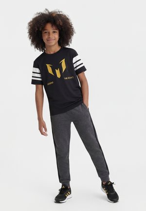 Chłopiec z kręconymi włosami, ubrany w czarny T-shirt ze złotym nadrukiem i napisem, szare spodnie dresowe i czarne trampki, stojący z jedną ręką w kieszeni.