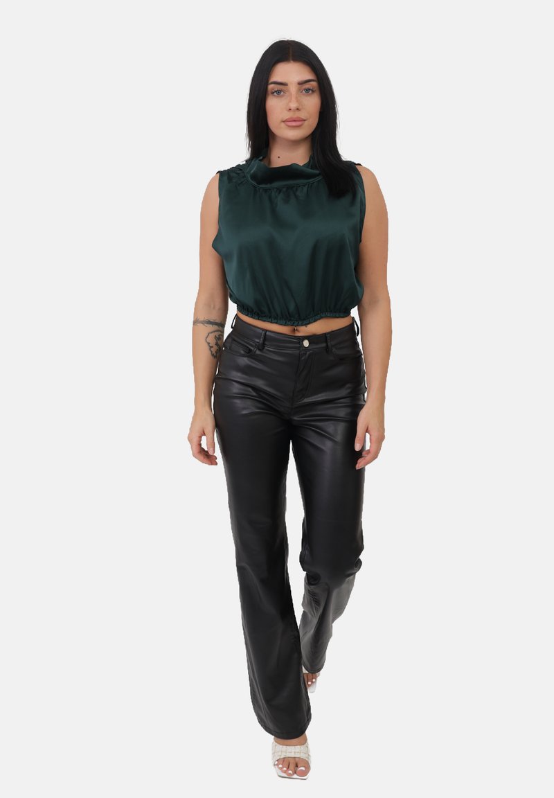 Kobieta idąca do przodu w ciemnozielonym satynowym topie bez rękawów, czarnych skórzanych spodniach, białych sandałach na obcasie i widocznym tatuażu na ramieniu.