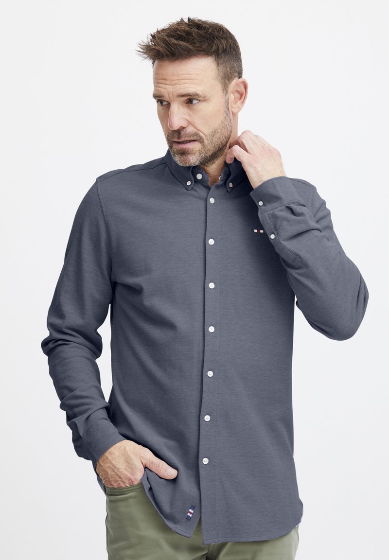 Chemise à manches longues en tissu gris foncé, avec des boutons blancs, un col boutonné et un petit accent de marque au niveau de l'ourlet.