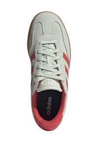 Ljust grå mockasneaker med röda ränder, platta snören, rundad tå och gummisulor. "adidas"-logotyp tryckt på plösen och innersulan.