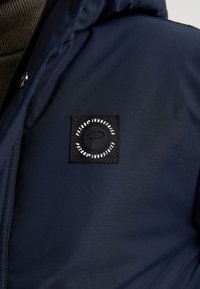 Giacca blu navy con una toppa quadrata nera con logo circolare, texture liscia, chiusura con zip e dettaglio del colletto a coste.