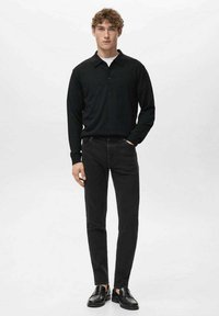 Polo de manga larga negro con cuello, combinado con vaqueros ajustados negros y mocasines negros, mostrado en un modelo masculino sobre un fondo blanco.