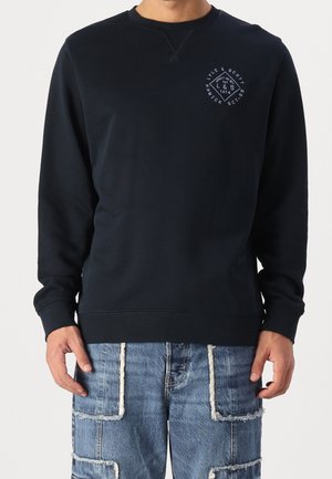 Mand iført en sort langærmet crewneck sweatshirt med lille hvidt logo på brystet og blå patchwork-jeans med frynsete kanter.