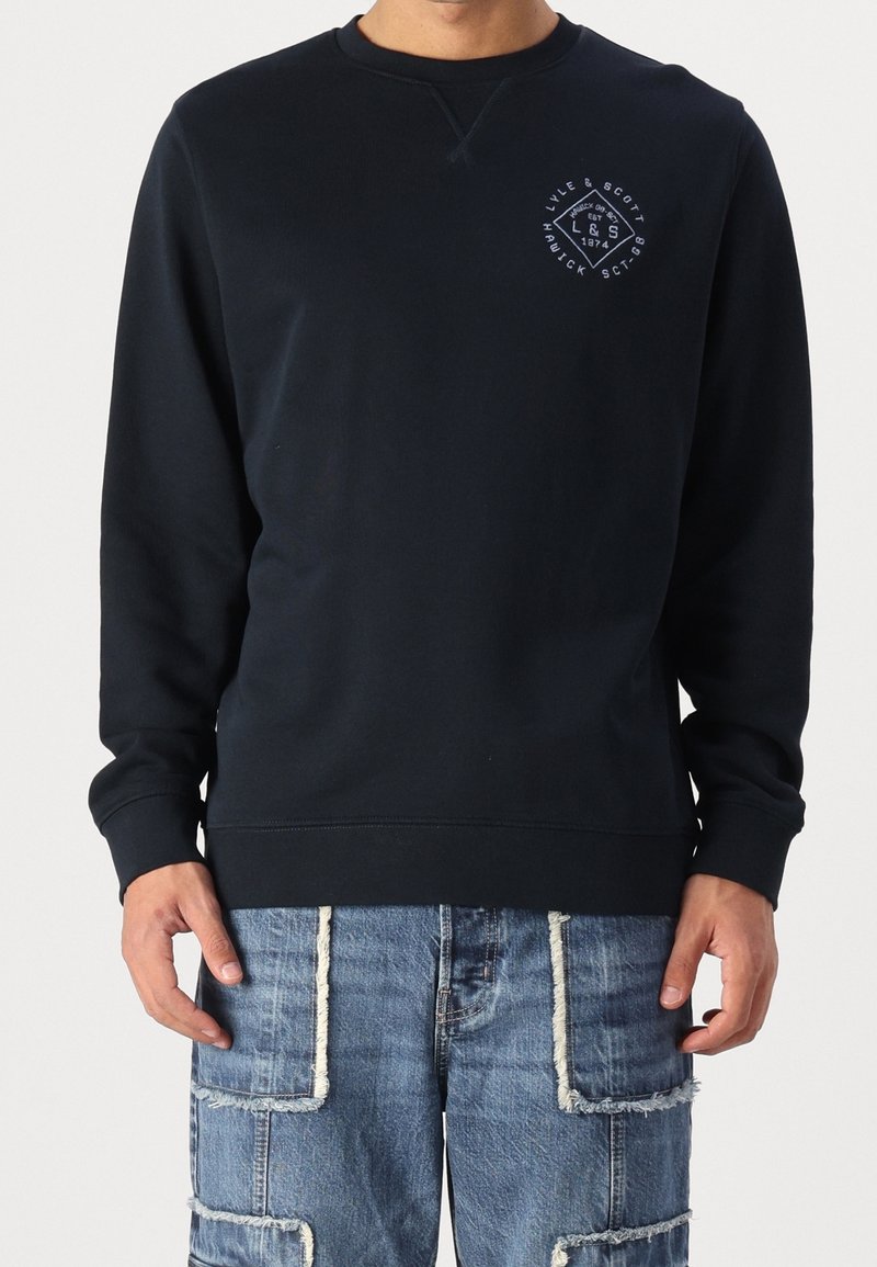 Homme portant un sweat-shirt noir à manches longues à col rond avec un petit logo blanc sur la poitrine et un jean patchwork bleu aux bords effilochés.