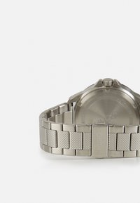 Orologio in acciaio inossidabile con finitura spazzolata e bracciale testurizzato. Fondello rotondo visibile, inciso con il logo del marchio e i dettagli.