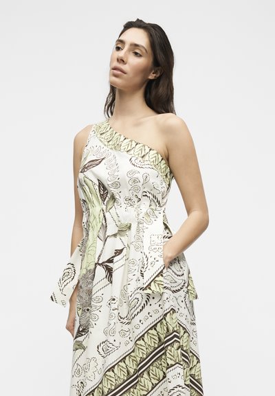 Γυναίκα που φοράει λευκό φόρεμα με έναν ώμο με πράσινα και καφέ αφηρημένα βοτανικά μοτίβα, όρθια με το ένα χέρι στην τσέπη.